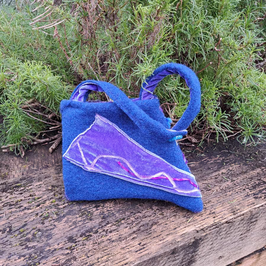 Blue felt bag with a triangular purple and lilac panel across the front, standing on weathered wood